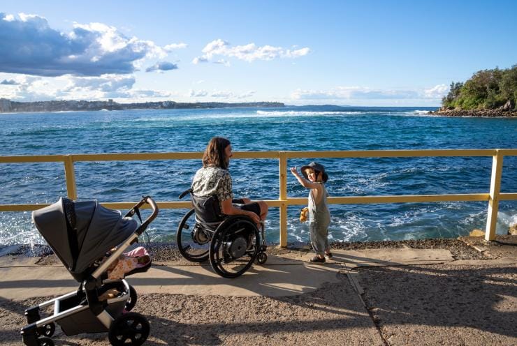 Một em nhỏ và một người đàn ông sử dụng xe lăn bên bờ biển tại Vùng Manly, Thành phố Sydney, Tiểu bang New South Wales © Tourism Australia