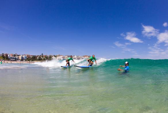 Một nhóm người đang học lướt sóng trên những con sóng màu xanh ngọc cùng Let's Go Surfing, Bãi biển Bondi, Tiểu bang New South Wales © Mikala Wilbow