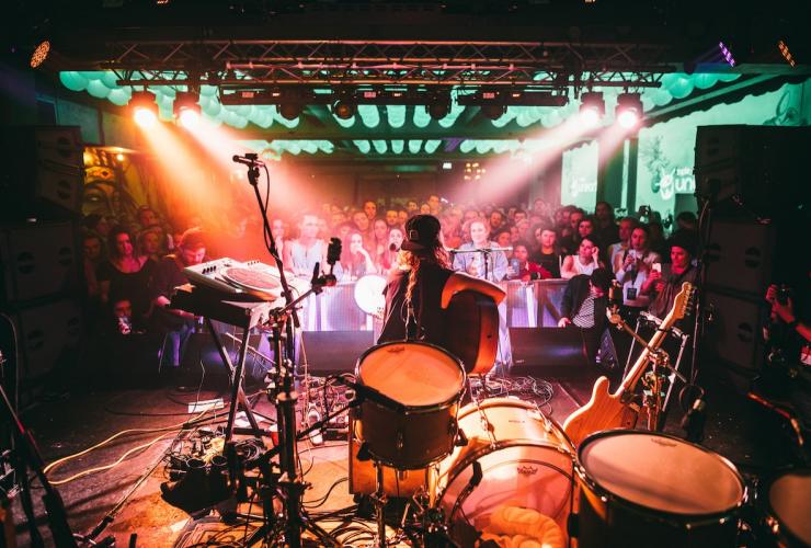 A person sitting on a stool playing a guitar among a stage filled with drums and microphones with a crowd of people watching in front of them during Bigsound, Fortitude Valley, Brisbane, Queensland © Stills in Time