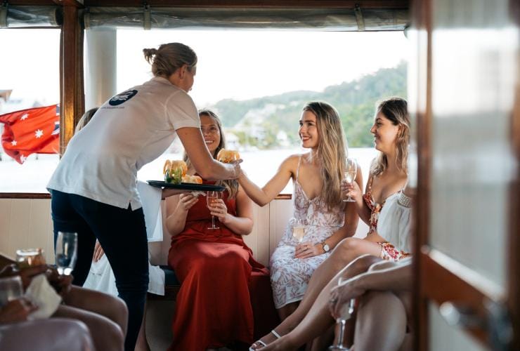 A waiter serving prawns and sparkling wine to a group of people sitting beside a window on a boat at Noosa Eat & Drink Festival, Noosa, Queensland © Tourism and Events Queensland