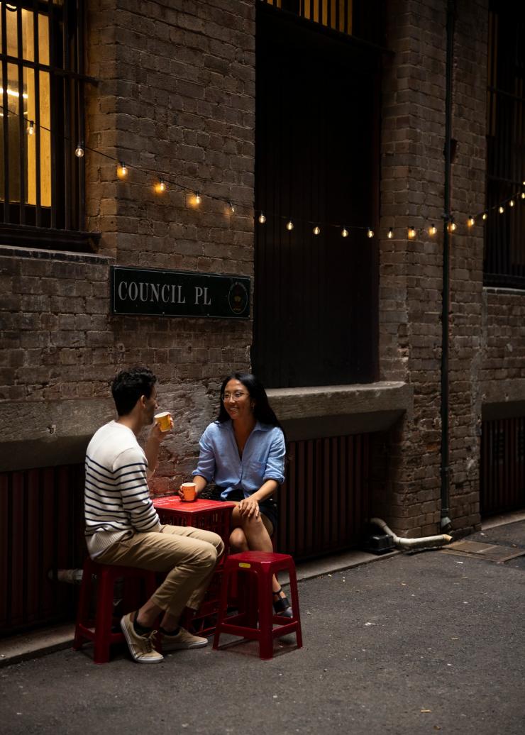 A man and woman seated in a laneway with a decorative string of lights above them while drinking cocktails at Cantina OK!, Sydney CBD, New South Wales © Destination NSW