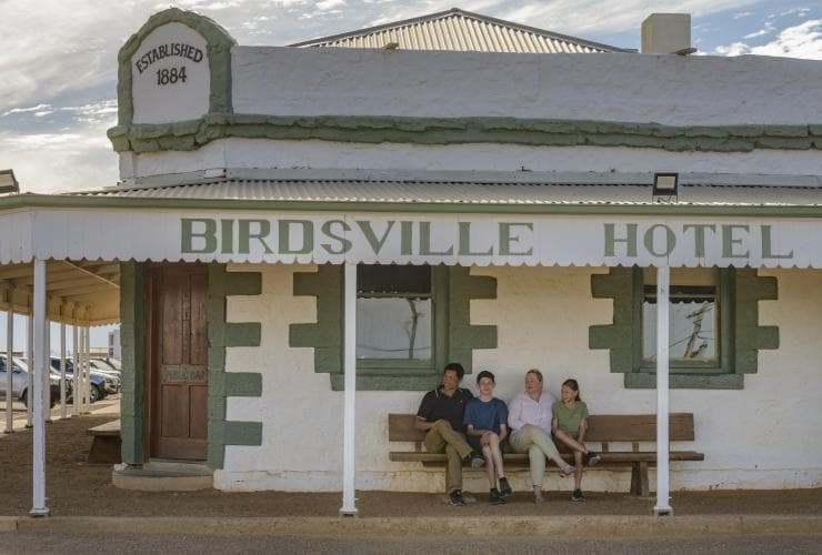 The exterior of a green and white historic pub, with a family of four sitting on a chair beneath the venue's awning at Birdsville Hotel, Queensland © Tourism and Events Queensland