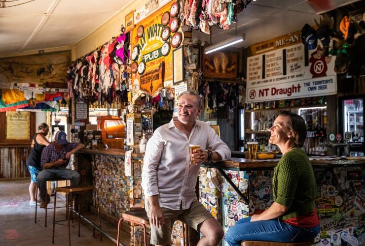 Two people sitting at a bar adorned in trinkets while enjoying schooners of beer at the Daly Waters Pub, Daly Waters, Northern Territory © Tourism NT/Helen Orr