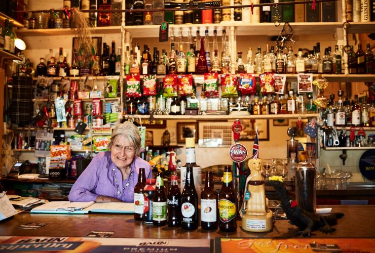 An elderly bartender smiling behind a bar while surrounded by bottles of alcohol at Grand Hotel Kookynie, Kookynie, Western Australia © Tourism Western Australia