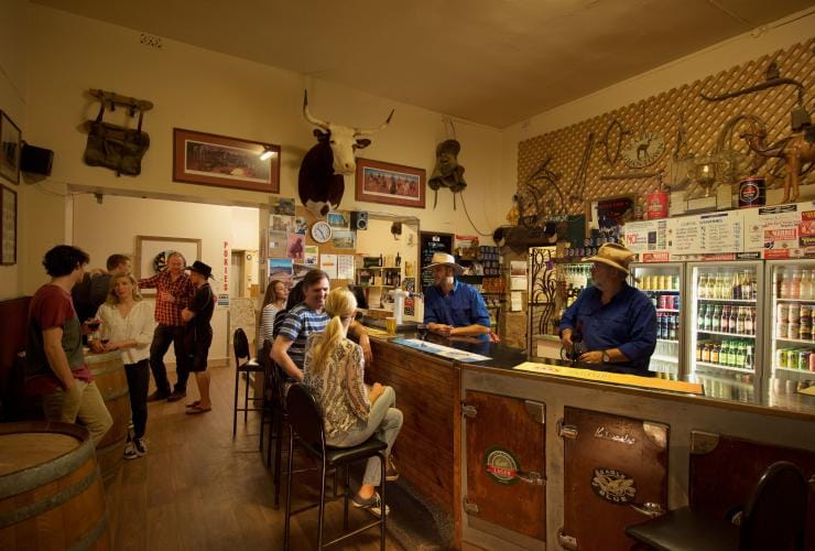 Two bartenders wearing cowboy hats and serving beer to a small crowd of people inside a quaint venue at the Marree Hotel, Flinders Ranges & Outback, South Australia © South Australian Tourism Commission