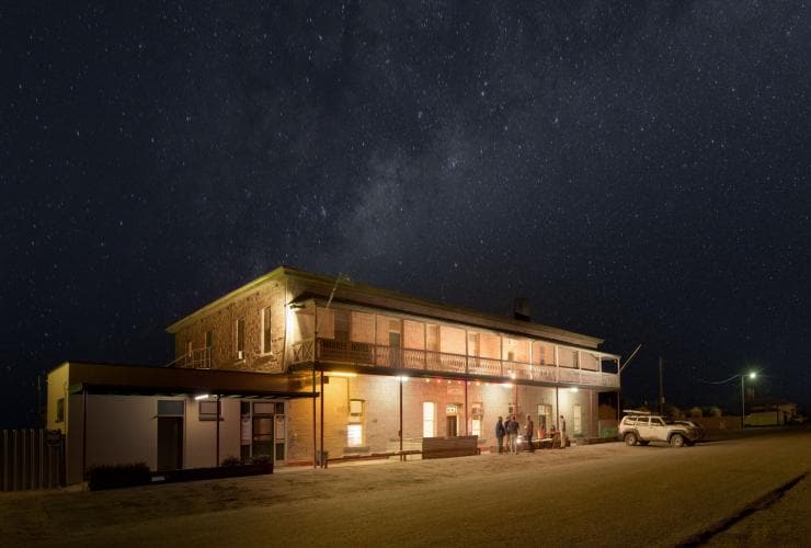 People standing in a circle enjoying drinks outside a venue at night beneath the stars at the Marree Hotel, Flinders Ranges & Outback, South Australia © South Australian Tourism Commission