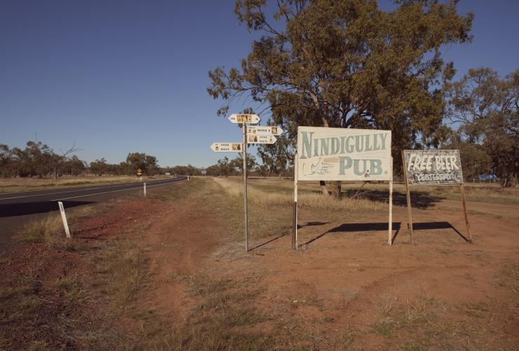 A large, vintage sign in between two smaller signs amid bushland on the side of a winding road at Nindigully Pub, Nindigully, Queensland © Tourism and Events Queensland 