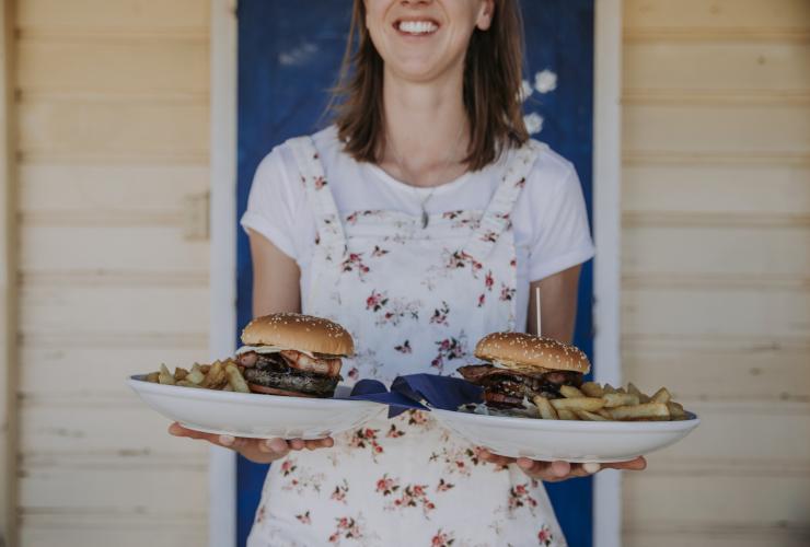 A person holding two plates with burgers and chips while smiling in front of a blue door at the Blue Heeler Hotel, Kynuna, Queensland © Tourism and Events Queensland 