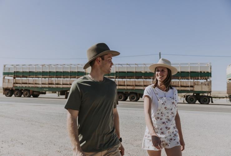 A man and woman walking on a sandy outback plain with a large truck behind them at the Blue Heeler Hotel, Kynuna, Queensland © Tourism and Events Queensland