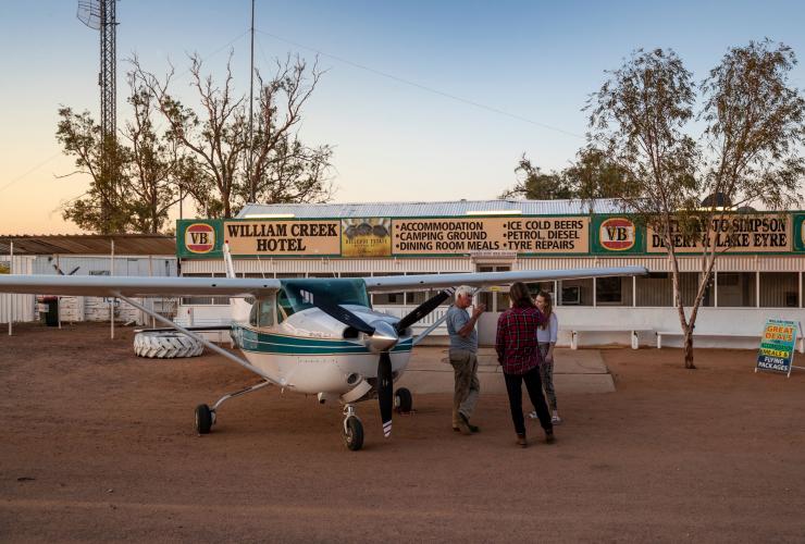 Three people standing beside a small plane parked outside a building with signs reading William Creek Hotel, Flinders Ranges & Outback, South Australia © South Australian Tourism Commission