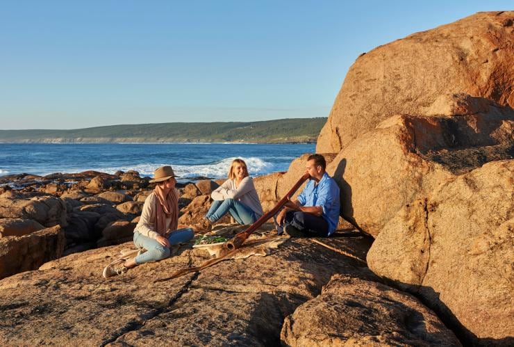 A person playing a didgeridoo with two people seated beside them on a rocky coastline with Koomal Dreaming, Margaret River, Western Australia © Frances Andrijich 