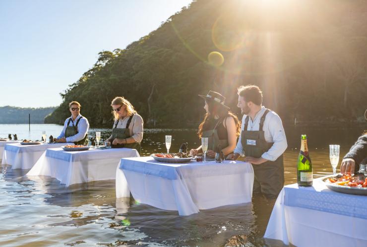 People standing in a river wearing waders at white tables with plates of seafood and sparkling wine with Sydney Oyster Farm Tours, Mooney Mooney, New South Wales © Tourism Australia