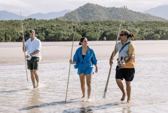 Hai du khách cầm xiên và cua bùn đi bộ qua một bãi bùn cùng hướng dẫn viên Thổ dân địa phương, Walkabout Cultural Adventures, Hẻm núi Mossman, Vùng Nhiệt đới Bắc Queensland © Tourism Australia