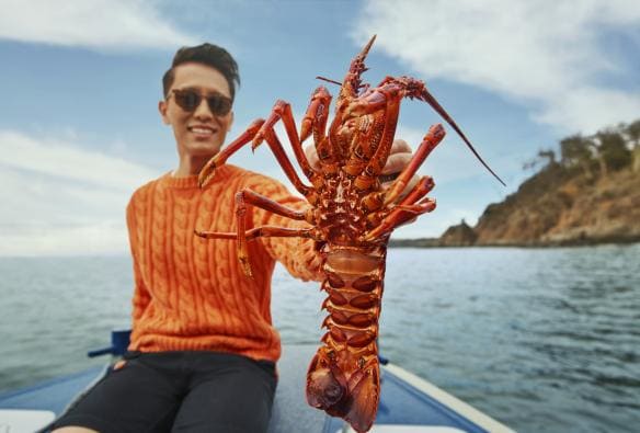 Một người ngồi trên thuyền, tay cầm tôm hùm hướng về phía máy ảnh, Seafood Seduction, Pennicott Wilderness Journeys, Đảo Bruny, Tiểu bang Tasmania © Tourism Australia