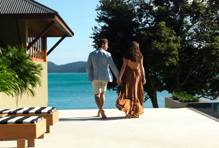 Couple holding hands walking past beach chairs towards the ocean at qualia, Hamilton Island, Queensland © Jason Loucas Photography