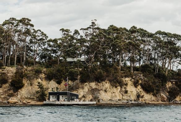 View across the calm blue ocean towards a seaside cottage beneath small rocky cliffs and native trees on Satellite Island, Tasmania © Adam Gibson