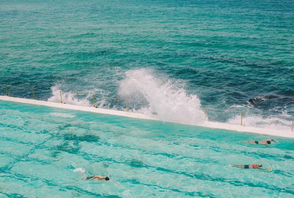 People swimming laps in the clear blue water of Bondi Icebergs Pool as the ocean’s waves crash against the side in Bondi, New South Wales © Tourism Australia