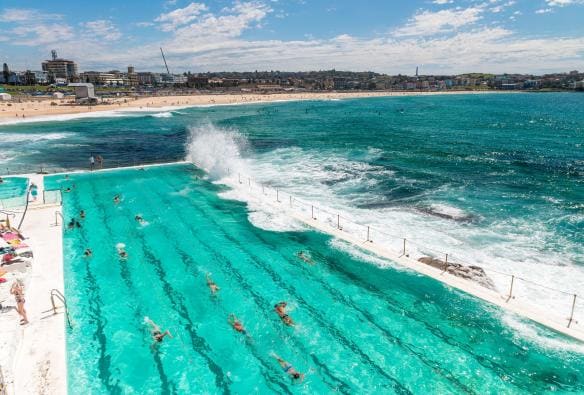View over people swimming laps in the turquoise water of Bondi Icebergs Pool as waves crash into the pool, with Bondi beach in the background in Sydney, New South Wales © Destination NSW