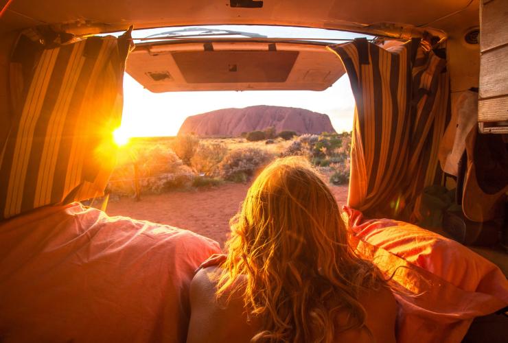A person lying in the back of a van with the doors opened giving way to views of Uluru during a bright golden sunrise, Uluru-Kata Tjuta National Park, Northern Territory © Mitchell Cox /Tourism NT