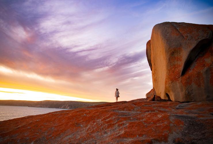 A woman walking beside an orange lichen-covered cliff face overlooking the ocean during sunrise with the Remarkable Rocks beside her, Kangaroo Island, South Australia © Tourism Australia