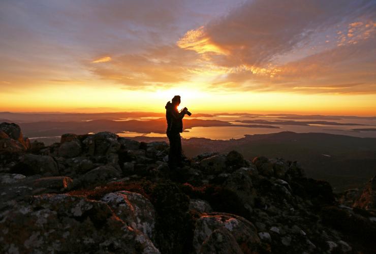 A person standing on rocks at the peak of kunanyi/Mt Wellington, looking into their camera as the sun shines around them across views of Hobart and surrounds in Tasmania © Tourism Australia