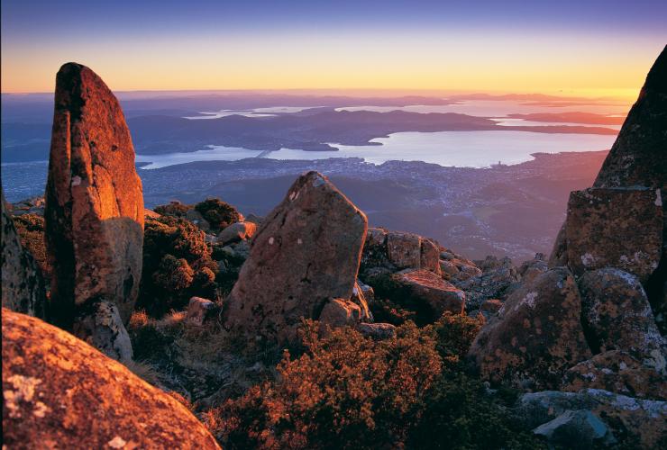 Rocks in the foreground on the summit of kunanyi/Mt Wellington glow red as the sunrise sweeps over views of Hobart and the ocean beyond in Tasmania © Tourism Tasmania / Garry Moore