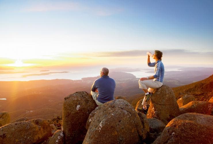 A couple sitting and relaxing on rocks at the peak of kunanyi/Mt Wellington as the sunrise casts an orange glow over Hobart below, Tasmania © Tourism Tasmania / Glenn Gibson