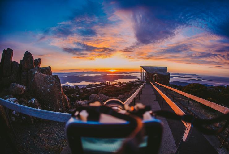A camera in the foreground capturing an image of a bright blue and orange sunrise over Hobart from kunanyi/Mt Wellington, Hobart, Tasmania © Tourism Australia