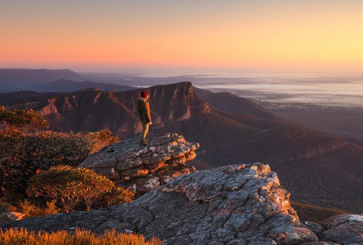 A person standing on the rocky peak of Mount William overlooking mountains, bushland and waterways during sunrise, Grampians National Park, Victoria © Parks Victoria/Matt Donovan
