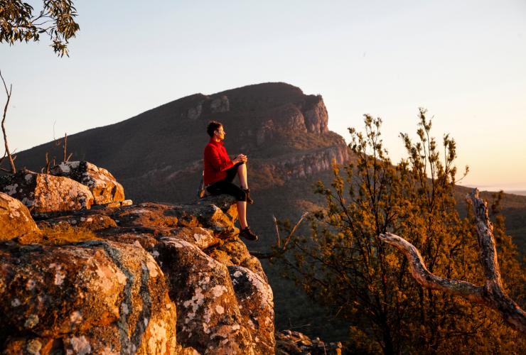 A person resting on a rock surrounded by wilderness as the sun creates an orange hue on their surroundings along the Picaninny (Bainggug) Walk, Grampians National Park, Victoria © Visit Victoria