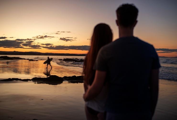 A couple standing in the foreground, watching a surfer run across the sand toward the ocean during sunrise at Snapper Rocks, Gold Coast, Queensland © Tourism and Events Queensland