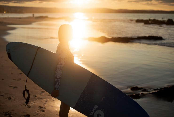 A surfer standing on the water’s edge holding their surfboard with the sun rising behind them at Snapper Rocks, Gold Coast, Queensland © Tourism and Events Queensland