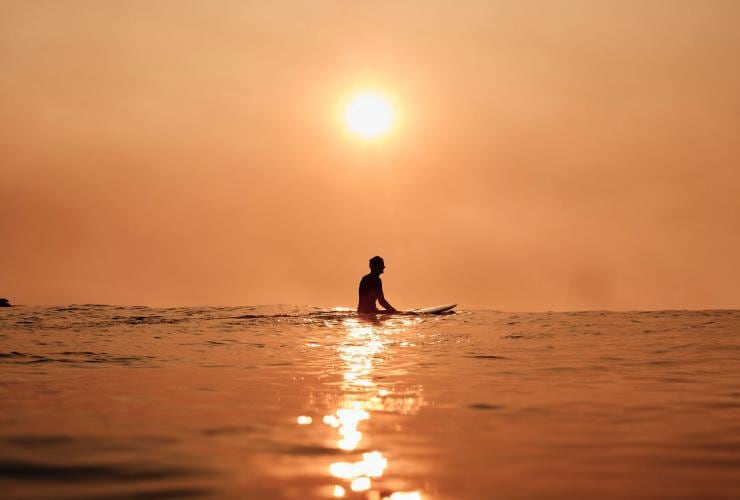 A person sitting on a surfboard beneath the sun glowing orange in the early morning at Bondi Beach, Sydney, New South Wales © Tourism Australia