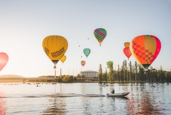 A group of colourful Hot Air Balloons floating above a boat and reflecting on the water of Lake Burley Griffin, Canberra, Australian Capital Territory © EventsACT