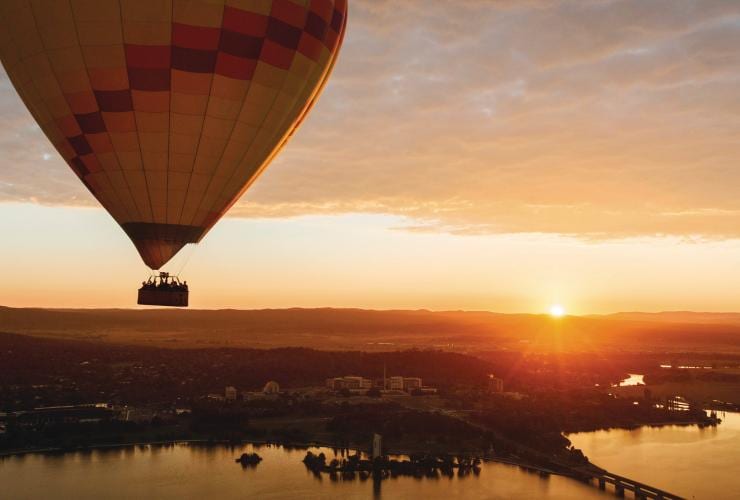 A hot air balloon floating above Canberra and Lake Burley Griffin as the sun rises on the horizon during Canberra Balloon Spectacular, Australian Capital Territory © Visit Canberra