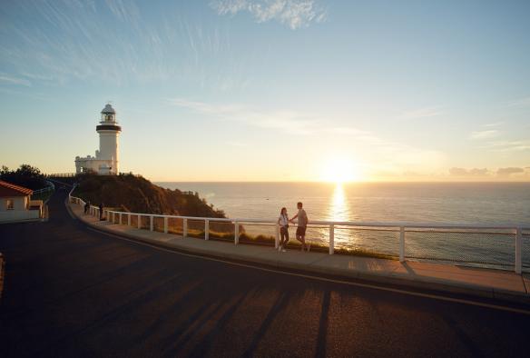 A couple leaning against a railing overlooking the ocean during sunrise with the Cape Byron Lighthouse to their left, Byron Bay, New South Wales © Hugh Stewart/Destination NSW