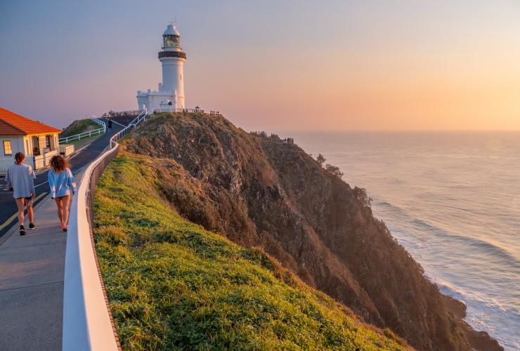 Two people walking along a path toward Cape Byron Bay Lighthouse with a grassy hill leading down to the ocean beside them, Byron Bay, New South Wales © Tourism Australia