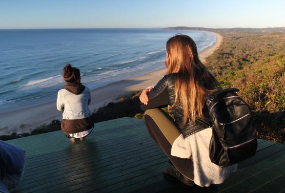 Two people sitting on a platform overlooking the lush green and white sand coast from Cape Byron Lighthouse, Byron Bay, New South Wales © Destination NSW