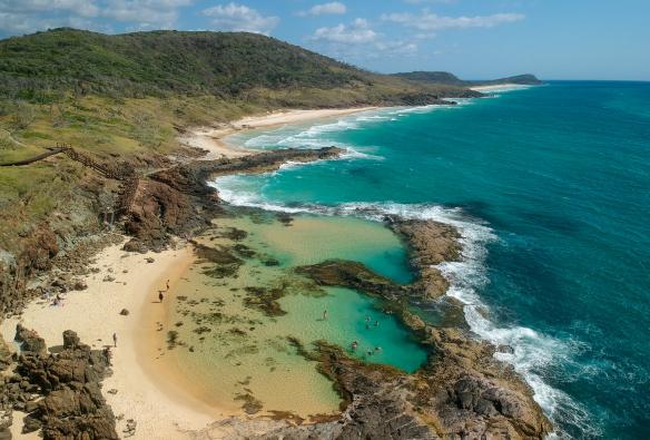 Ảnh chụp từ trên cao cảnh nhóm du khách đang bơi trong các hồ nước tự nhiên Champagne Pools với những con sóng cuộn tràn vào mỏm đá ngăn cách giữa hồ nước và đại dương ở đảo K’gari, Tiểu bang Queensland © Tourism Australia