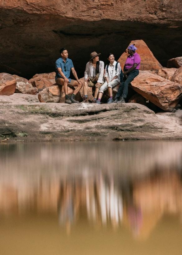 Một nhóm du khách đang ngồi cùng hướng dẫn viên Thổ dân địa phương từ Kingfisher Tours bên dưới khối đá lớn với dòng nước phía trước tại Vườn quốc gia Purnululu, Tiểu bang Tây Úc © Tourism Western Australia