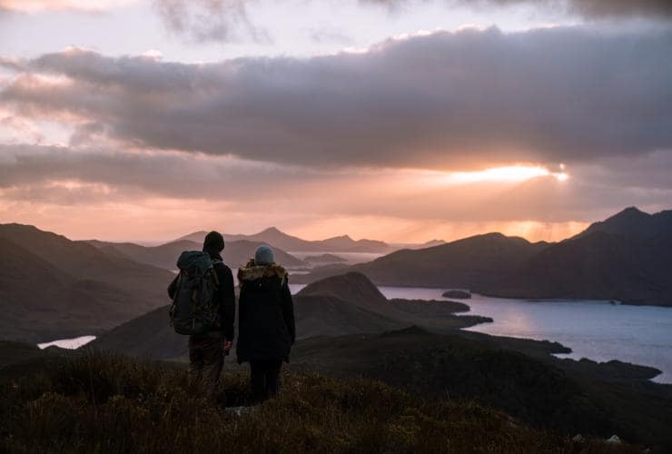 Man and woman with backpacks on a hike looking  at the view of Bathurst Harbour at sunset, Southwest National Park, Tasmania © Jason Charles Hill