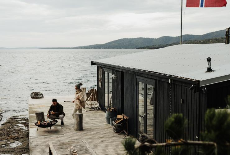 Man crouching beside a fire pit beside waterfront accommodation while woman stands with a marshmallow ready to toast on Satellite Island, Tasmania © Adam Gibson
