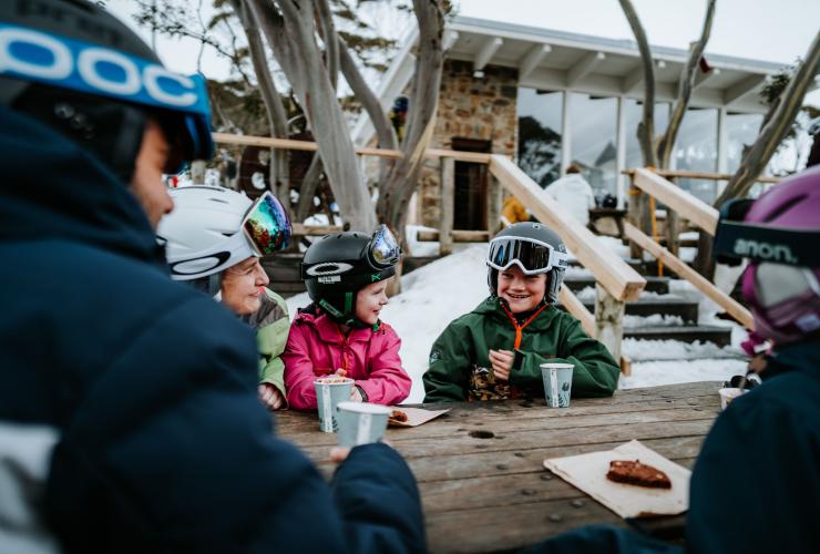 Một gia đình đang vui cười thưởng thức bánh quy và sô-cô-la nóng tại bàn dã ngoại giữa tuyết, Núi Hotham, High Country, Victoria © Tourism North East