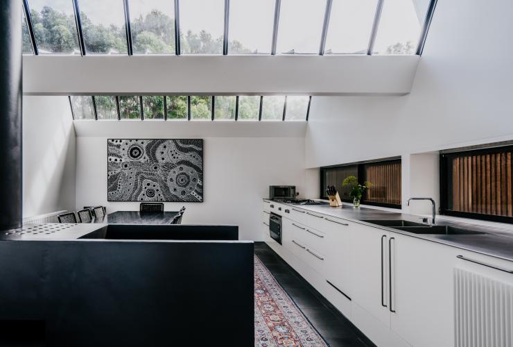A large, modern kitchen and dining area with an Aboriginal artwork on the wall and windows lining the roof and drenching the room in natural light at Alkina Lodge, Wattle Hill, Victoria © Alkina Lodge
