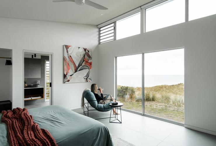 A person sitting on a lounge chair sipping wine while looking out through large windows towards a rugged coastline at Kittawa Lodge, King Island, Tasmania © Kittawa Lodge/Oscar Sloane
