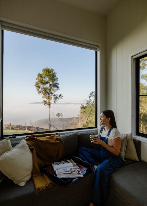 Một người đang ngồi trên ghế dài, ngắm nhìn khung cảnh vùng nông thôn ngoài cửa sổ, Baya Tiny House, Scenic Rim, Tiểu bang Queensland © Tourism and Events Queensland