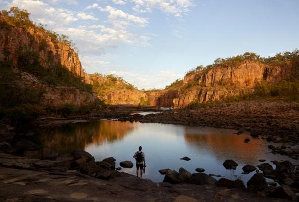 Hiker pausing near a still lake surrounded by red cliffs in Nitmiluk National Park, Northern Territory © Tourism NT/Matt Cherubino
