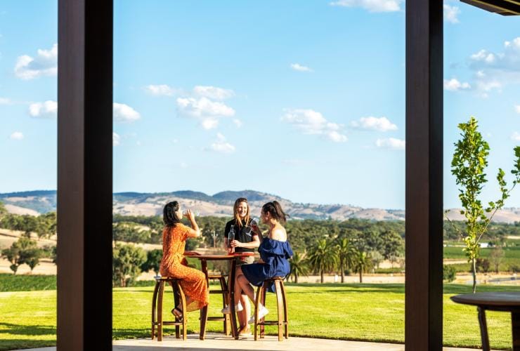 Three women laugh and sip wine at an outdoor table at © Tourism Australia