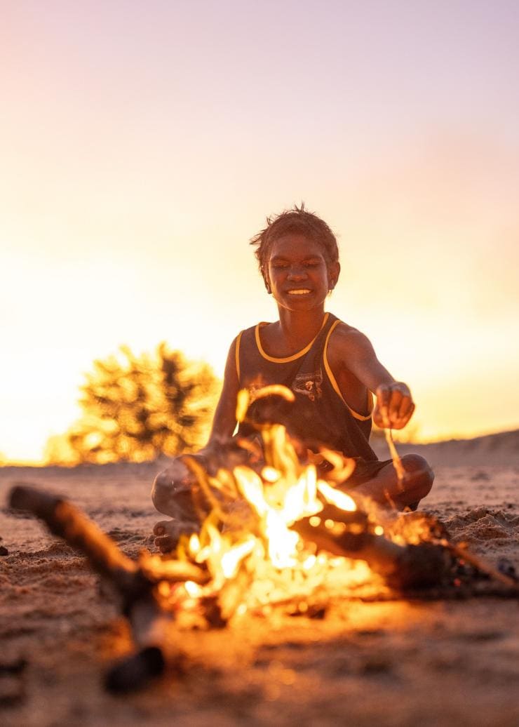A young local boy sits behind a campfire on the beach at Tarntipi Bush Camp, Tiwi Islands, Northern Territory © Tourism NT/Helen Orr