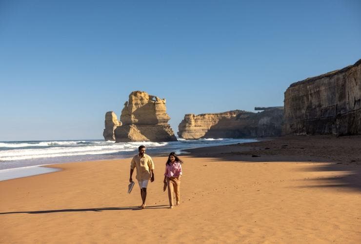 A couple walks along the golden sand with rock formations behind them at the Twelve Apostles, Great Ocean Road, Victoria © Tourism Australia